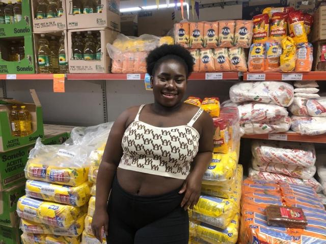 Dilay, con la pelle scura, i capelli ricci e corti e un sorriso, indossa una canotta bianca con motivi e pantaloni neri, ed è in piedi in un supermercato con sacchetti gialli e arancioni..