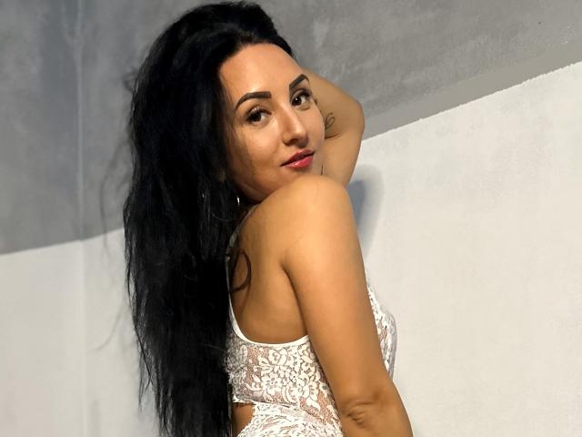 BelleAndra mit langem, schwarzem Haar, die über ihre Schulter schaut, trägt ein weißes Spitzen-Bralette, das ihre Brüste zeigt und hat ein kleines Tattoo auf ihrem Oberarm..