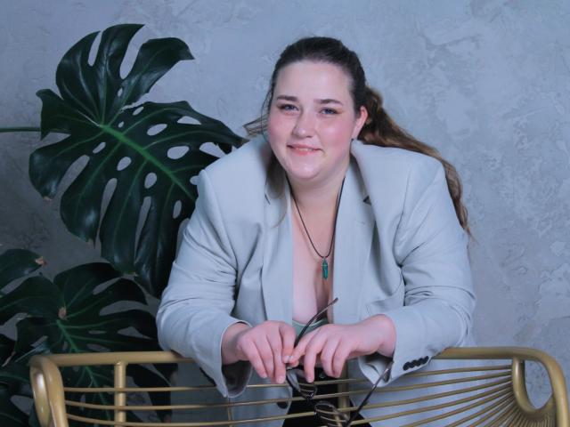 DaniellaGraceZZ with brown hair, gray blazer, and green necklace, leaning on gold chair, smiling, hands on glass, large green monstera plant behind her, blue-gray wall.