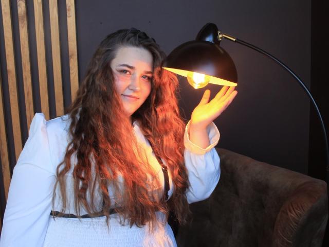 DaniellaGraceZZ with long wavy brown hair, white shirt, holding black lamp, light shining on her face, smiling, brown couch in background, warm light, soft shadows, relaxed vibe.