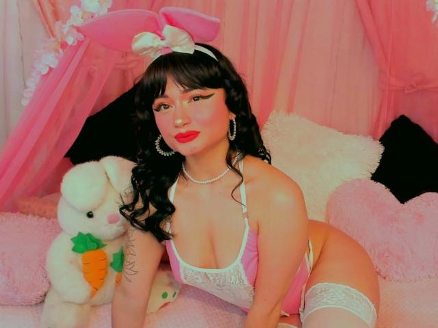 AnnieGrey with pink bunny ears, white lace lingerie, and thigh-highs, sitting on pink pillows, holding a white stuffed bunny with a carrot, showing her tits and a tattoo.