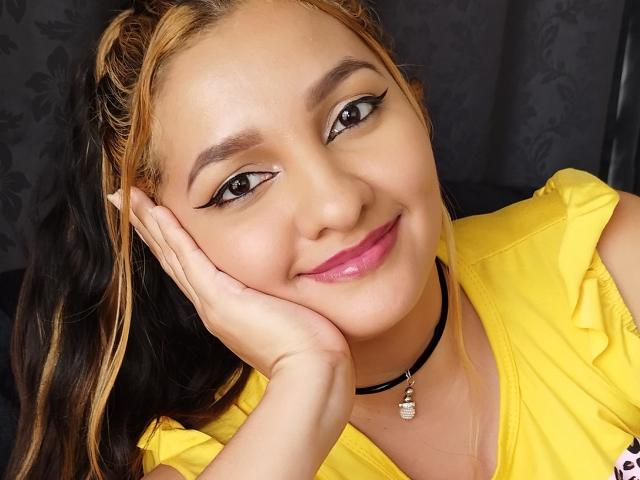 GisellHotty, com cabelo loiro e preto, vestindo uma camisa amarela, um colar preto e batom rosa, sorrindo com a mão no rosto, delineador realçando seus olhos, e um rosto adorável..