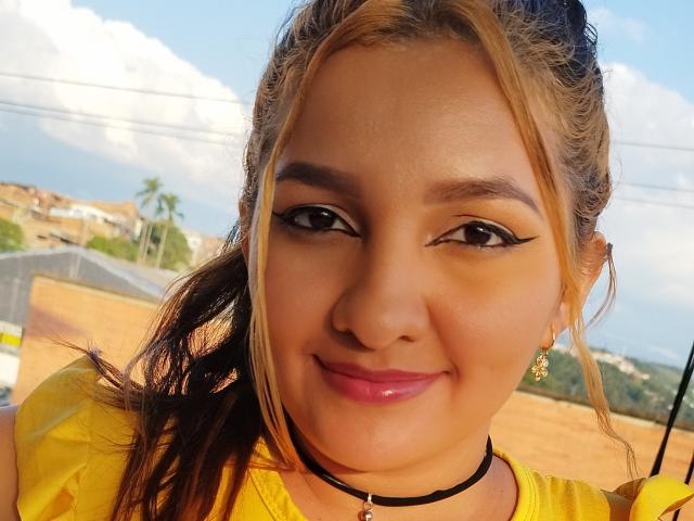 GisellHotty with light brown skin, blonde highlights, black eyeliner, pink lipstick, yellow top, black choker, smiling, brown eyes, small gold earrings, ponytail, outdoor, sunny, clear sky.