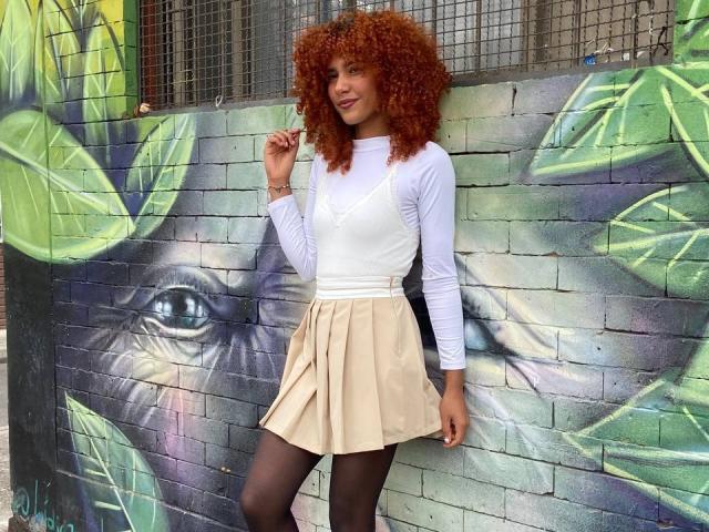 CinthiaWalker with curly red hair, white long-sleeve top, beige pleated skirt, black tights, standing against graffiti wall, smiling, hand in hair, showing her ass subtly with skirt.
