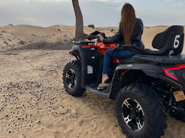 BeataAngel with long brown hair, black leather jacket, and blue jeans, sits on a black and orange ATV in a sandy desert, wearing sandals, facing away, holding a phone.