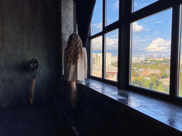 BeataAngel with long, wavy blonde hair, standing by a large window, wearing a white dress, looking outside, with a vintage camera on a tripod in a dimly lit room.