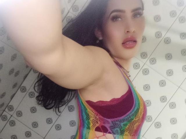 Ein Selfie von ElectraHottyX, auf dem ihr Arm erhoben ist. Sie trägt ein bunter Mesh-Top, unter dem ein roter Spitzen-BH sichtbar ist. Sie hat schwarzes Haar, Make-up und pinke Lippen. Der Hintergrund ist ein Badezimmer mit Fliesen..