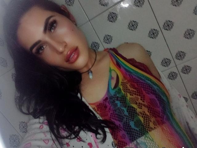 ElectraHottyX met lang zwart haar, draagt een kleurrijke regenboog top van netstof, heeft glanzende rode lippen en een schattige ketting, en laat een sexy gezicht en borsten zien in een badkamer..