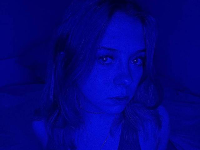 Foto com iluminação azul de ColdEmbrace, com cabelo loiro ondulado, olhando intensamente. Ela está usando um colar fino, seus olhos estão ligeiramente semicerrados e ela tem uma expressão sutilmente emburrada..