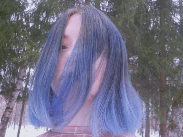 ColdEmbrace, con i capelli che sfumano dal blu al viola e che coprono il viso, mostrando parzialmente il naso e la bocca, in piedi in una foresta innevata con alberi verdi alti..