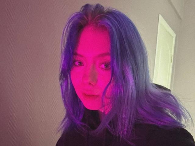 ColdEmbrace ha i capelli rosa e viola, un piercing al naso e indossa una maglietta nera, con un'espressione seria, in un selfie dai colori vivaci, illuminato di viola..