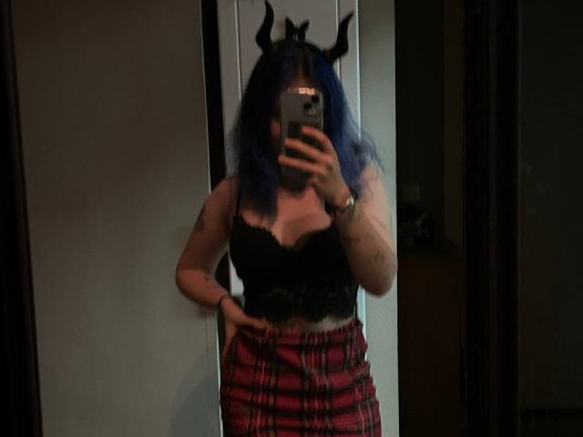 ColdEmbrace mit blauen Haaren, schwarzem Spitzenoberteil, rotem Karo-Rock und schwarzem Horn-Haarband, macht ein Selfie vor einem Spiegel und zeigt kleine Brüste und ein Tattoo am linken Arm..