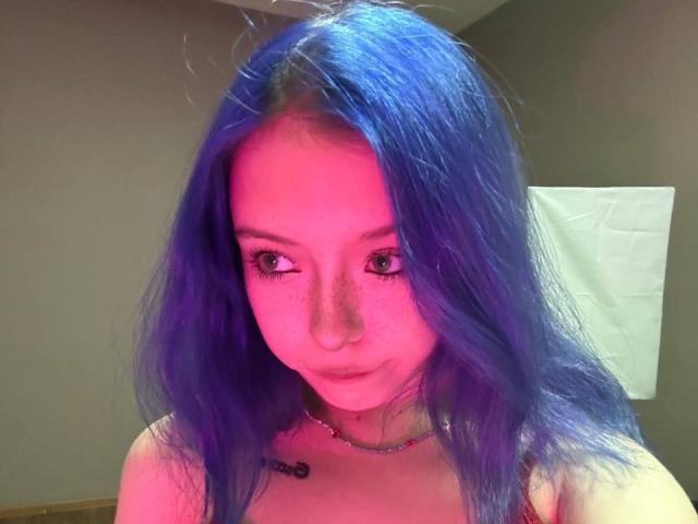 ColdEmbrace ha i capelli di un blu acceso, un'illuminazione rosa-viola e delle lentiggini. Sta guardando in basso con un leggero broncio, indossando una maglietta rossa e una collana. Il suo seno è piccolo..