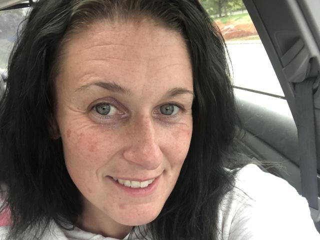 RDreamzCumTru tem pele clara, cabelo preto ondulado, olhos verdes, um leve sorriso e um pouco de vermelhidão nas bochechas, sentada em um carro vestindo uma camisa branca..
