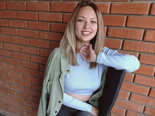 SallyVerda with blonde hair, smiling, wearing a green shirt over a blue crop top, sitting on a chair against a brick wall, hand near chin, looking cute.