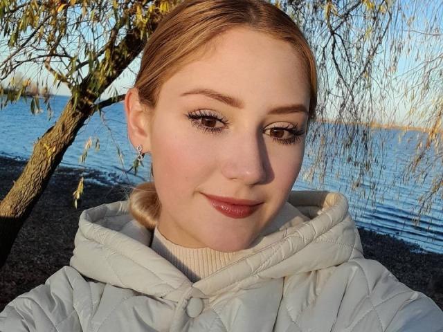 LaylaHott has pale skin, light brown hair, and wears a white quilted jacket. She's smiling with pink lipstick, brown eyes, and eyeliner. Background has a tree and water.