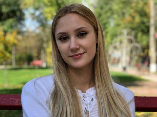 LaylaHott con lunghi capelli biondi, indossa una camicia bianca, sorride dolcemente, in piedi di fronte a una ringhiera di legno rossa, con uno sfondo sfocato di un parco verde..