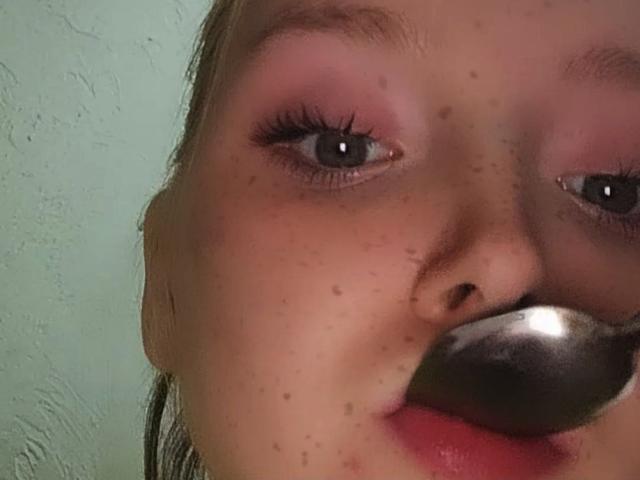 El rostro de LaPochka, con sombra de ojos rosa, ojos azules y un mordisque metálico brillante en la boca. Tiene pecas, labios rosados, y su nariz es parcialmente visible..
