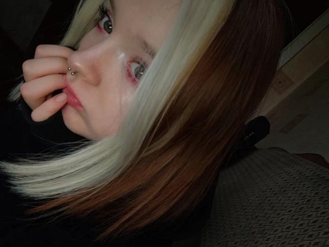 LaPochka, avec ses cheveux blonds et bruns, son piercing au nez et ses yeux roses, mordille son doigt, vêtue de noir, assise sur un siège gris à motifs. Sa main est près de sa bouche..