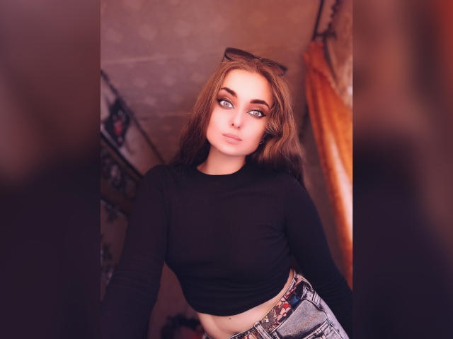KariMillie tiene el cabello castaño claro, ojos azules y una camiseta negra de manga larga que deja ver sus pequeños senos y una cinturilla estampada, con una ligera sonrisa y gafas en la cabeza..