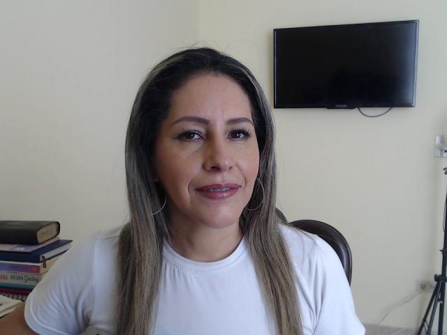 Carolina Ruiz a de longs cheveux bruns et lisses, porte une chemise blanche, des boucles d'oreilles cercles, et a un léger sourire, avec des fesses plates et de petites seins. Elle est assise devant un téléviseur fixé au mur..