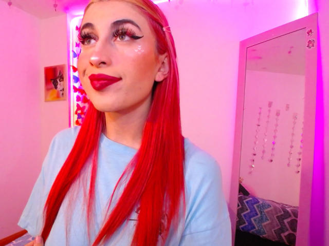SammyViolet with red hair, blue shirt, and red lipstick, showing off her tits in a pink-lit room, mirror with strings in background. Her ass is not visible.