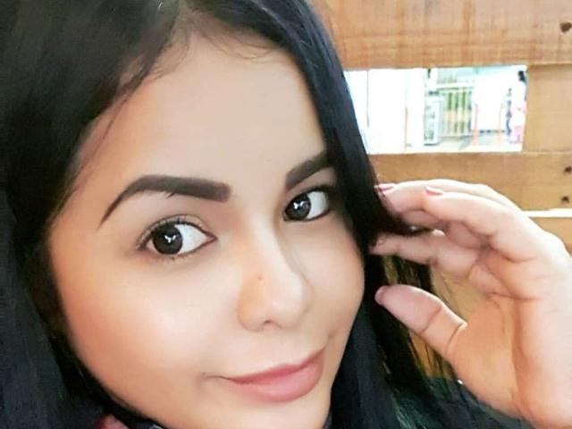 SmiaHot tiene una piel marrón media, cejas negras, ojos grandes marrones, cabello largo negro y una sonrisa sutil. Está tocándose el cabello con su mano derecha..