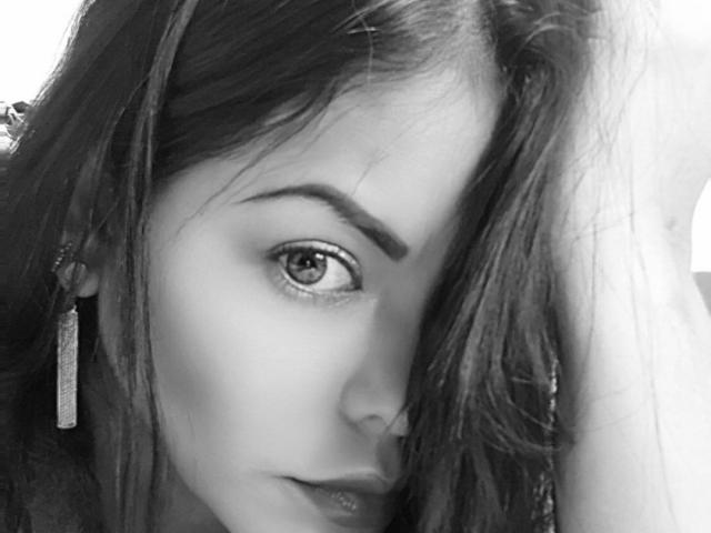 SmiaHot heeft lang, golvend haar dat één oog bedekt, draagt een enkele, bungelende oorbellen en heeft een lichtjes open lippen met een glanzende uitstraling. Zwart-wit portret, gefocust op haar oog..