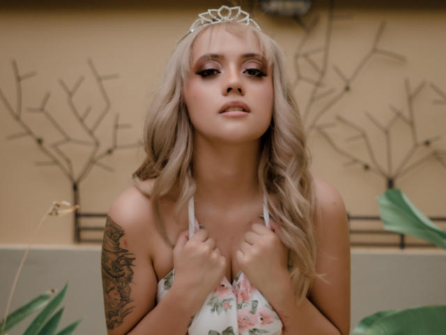 SabrinaCole, avec ses cheveux blonds ondulés, portant un haut à bretelles fleuri, tenant sa poitrine, une couronne sur la tête, un tatouage sur le bras gauche, regardant vers le bas, avec des feuilles vertes au premier plan..