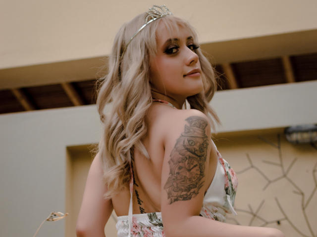SabrinaCole, com cabelos loiros ondulados, vestindo um vestido floral, mostrando uma tatuagem no ombro direito, olhando por cima do ombro, sorrindo, em uma luz suave, ao ar livre..