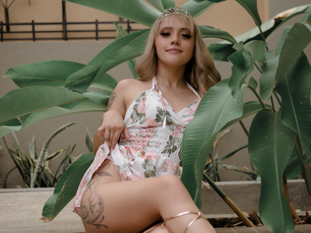 Sabrina Cole, con i capelli biondi, un vestito floreale e una tiara, seduta tra le foglie verdi, che mostra un tatuaggio sulla coscia destra, con la biancheria intima rosa visibile, e un sorriso rilassato..