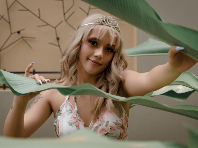 SabrinaCole, com cabelos loiros ondulados, vestindo um vestido floral e uma tiara, sorrindo entre folhas verdes, mostrando seus seios, esmalte azul nas unhas e uma tatuagem no braço direito..
