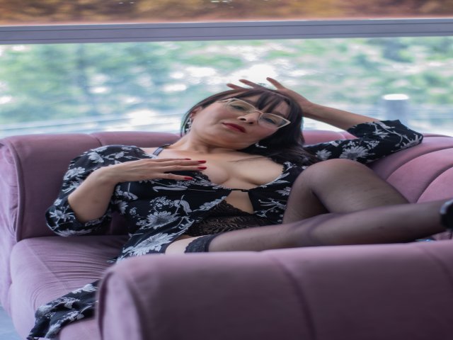 LexiLuna with glasses, black floral dress, and stockings, lounging on a pink chair, hand on head, other hand on her tits, legs crossed, red nails, looking relaxed.