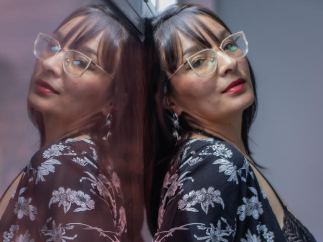 LexiLuna mit Brille, rotem Lippenstift und einem schwarzen, mit Blumenmuster versehenen Bademantel, wobei die Spiegelung ihren Rücken und ihr Gesicht zeigt. Sie hat ein süßes, leicht nerdiges Aussehen, mit glattem, braunem Haar..