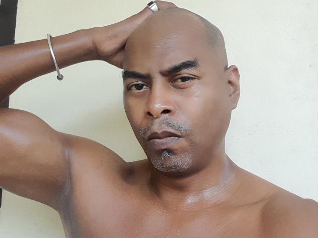Shiny-skinned DocLickALot with a bald head, dark eyebrows, and a small mustache, holding his head with a hand, wearing a silver ring and bracelet, looking serious.