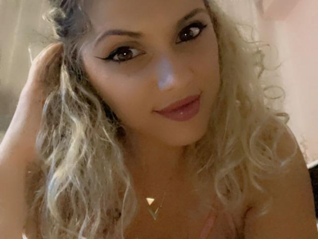 LucilleXSugar, con cabello rubio rizado, ojos marrones seductores y labios rosados, sonriendo ligeramente, tocándose el cabello con la mano derecha, llevando un collar de forma triangular, en una imagen con iluminación suave..