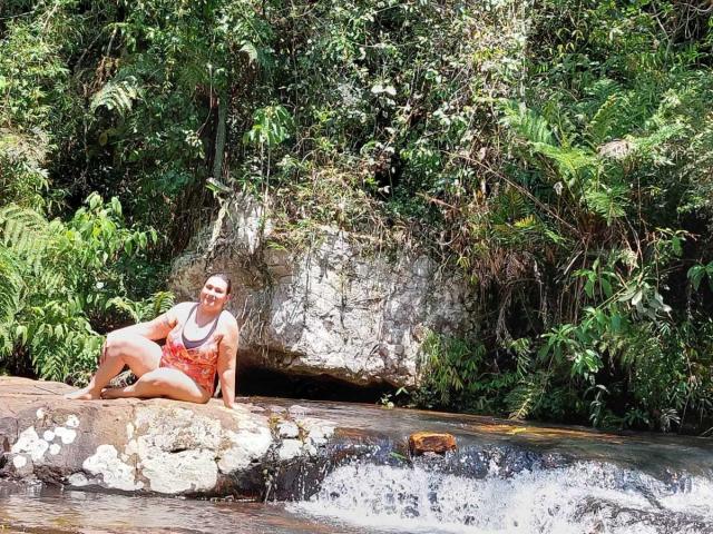 LadySelene, with her tan skin and red-orange tank top, sits on a rock by a small waterfall, her ass slightly raised, wearing black sandals in a lush forest.
