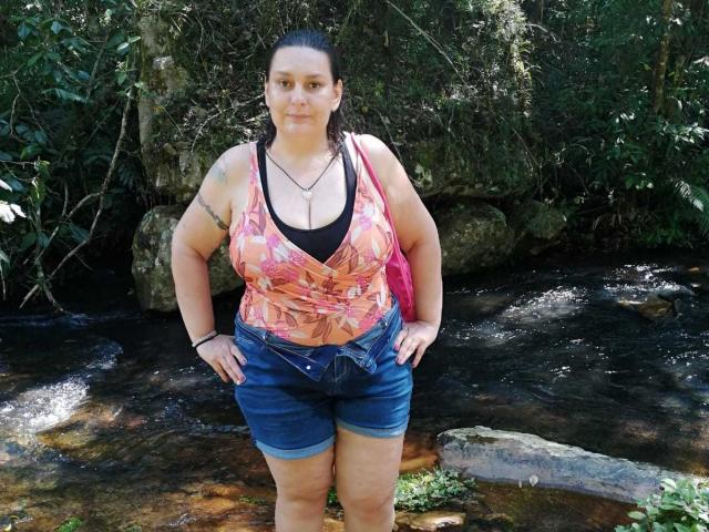 LadySelene with big tits, wearing a pink floral top, black bra, and blue shorts, standing in a creek, hands on hips, tattoo on right arm, black hair in a ponytail.