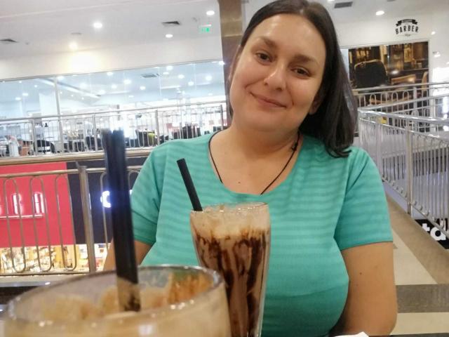LadySelene with medium boobs, wearing a teal shirt, smiling, holding a tall glass with black and white stripes, a black straw, in a brightly lit cafe.