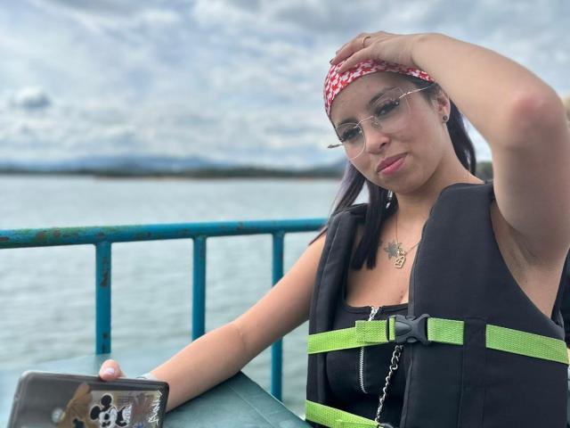 Samantha Morgan con gli occhiali, una bandana rossa, un gilet nero, cinghie verdi, che tiene un telefono, un tatuaggio sul petto, una mano sulla testa, acqua e una ringhiera blu sullo sfondo..