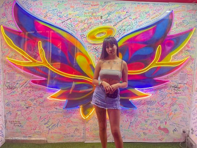 Samantha Morgan se tient debout, avec des ailes de papillon néon, vêtue d'un haut blanc moulant et d'une jupe en jean bleu usée, mettant en valeur ses seins et ses fesses, devant un mur coloré, décoré dans un style graffiti..