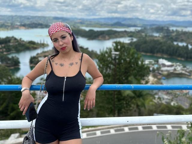 Samantha Morgan, con un busto de tamaño medio, lleva un pañuelo de cuadros rojos y blancos, un vestido negro con cremallera y está de pie junto a una barandilla azul y blanca, sosteniendo un bolso negro..