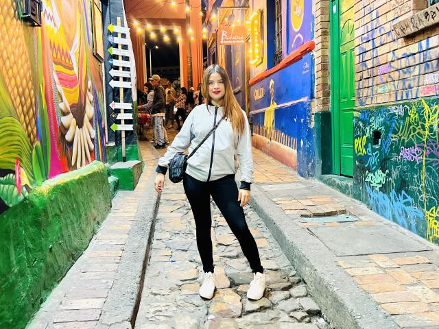 ShantalSummer has a cute ass, black pants, white sneakers, and a gray jacket, standing in a colorful, graffiti-filled alley with string lights and a small crowd in the background.
