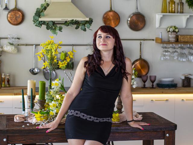 NinaFlawlees with red hair, black dress, and pink nails, leaning on a wooden table with yellow flowers and candles, showing her ass, in a kitchen.