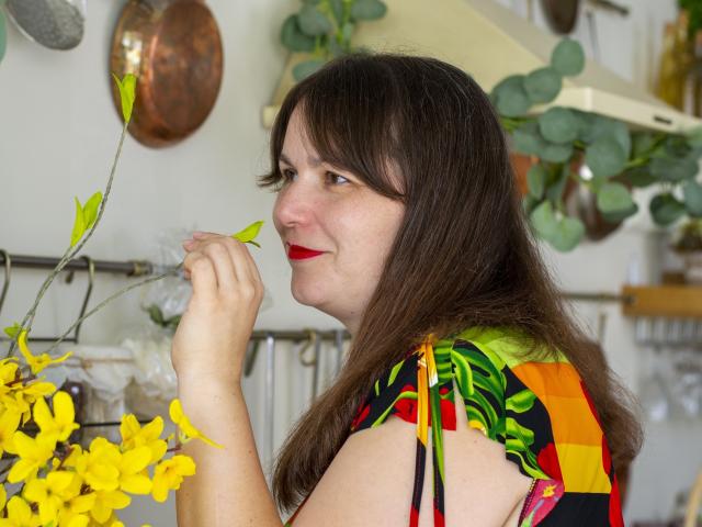 AliceInks with medium tits, brown hair, red lipstick, colorful dress, holding yellow flowers, smiling, white background, copper pots, green leaves, soft light, casual, relaxed, attractive.