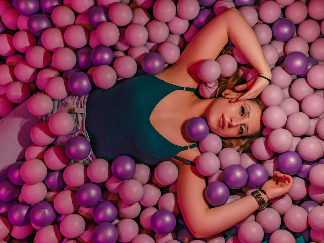 LaiaReyd liegt in einem violett-rosa Bällebad, trägt ein türkisfarbenes Tanktop, hat die Arme über dem Kopf und ein Armband am Handgelenk und schaut in die Kamera..