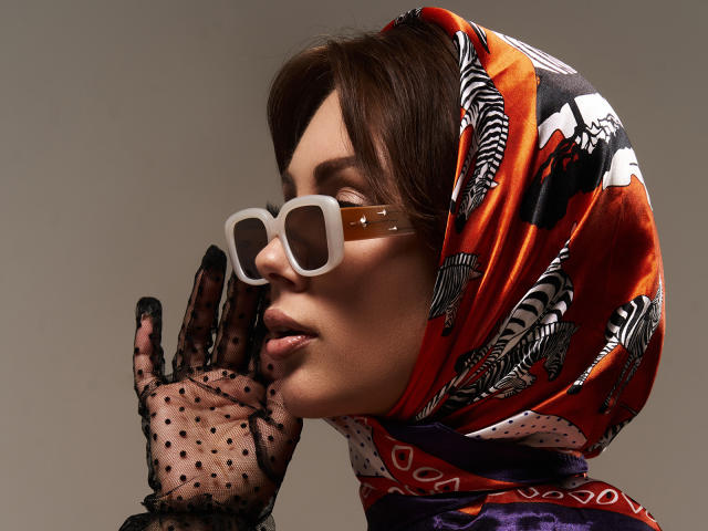 NinaTender, avec de grandes lunettes blanches, un foulard à motifs orange et rouge, des cheveux bruns, des gants noirs à pois, un bandage marron sur le nez, vue de profil, fond gris..