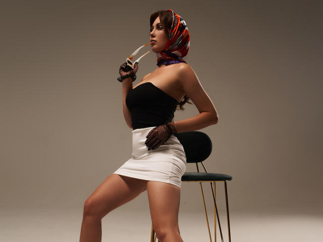 NinaTender with red headscarf, black strapless top, white mini-skirt, black gloves, holding cigarette, sitting on green chair, showing off her ass and taut legs.