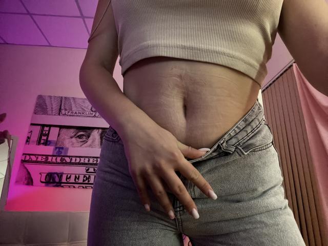 Destinis's belly button, hand in gray jeans, pulling down slightly, revealing a bit of white underwear, wearing a beige ribbed crop top, showing a small, soft, untoned belly.