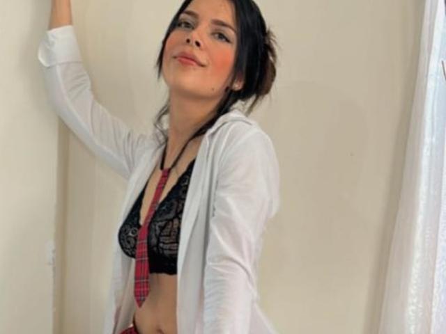 Sofia Rotavisky draagt een zwarte kanten bh, een rode geruite das en een open wit overhemd, staat tegen een muur, glimlacht en heeft haar arm omhoog, waarbij haar borsten zichtbaar zijn. Haar achterkant is niet te zien..